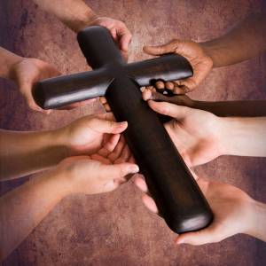 Several hands of people of different ethnicities hold a small wooden cross