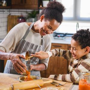 A little boy helps his mother in the kitchen by scooping food from a mixing bowl into a dish.